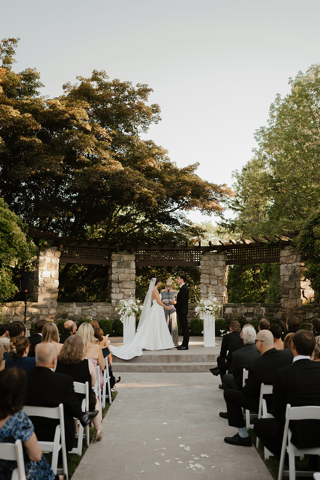Outdoor wedding ceremony with guests seated in white chairs watching the couple exchange vows at the stone altar.